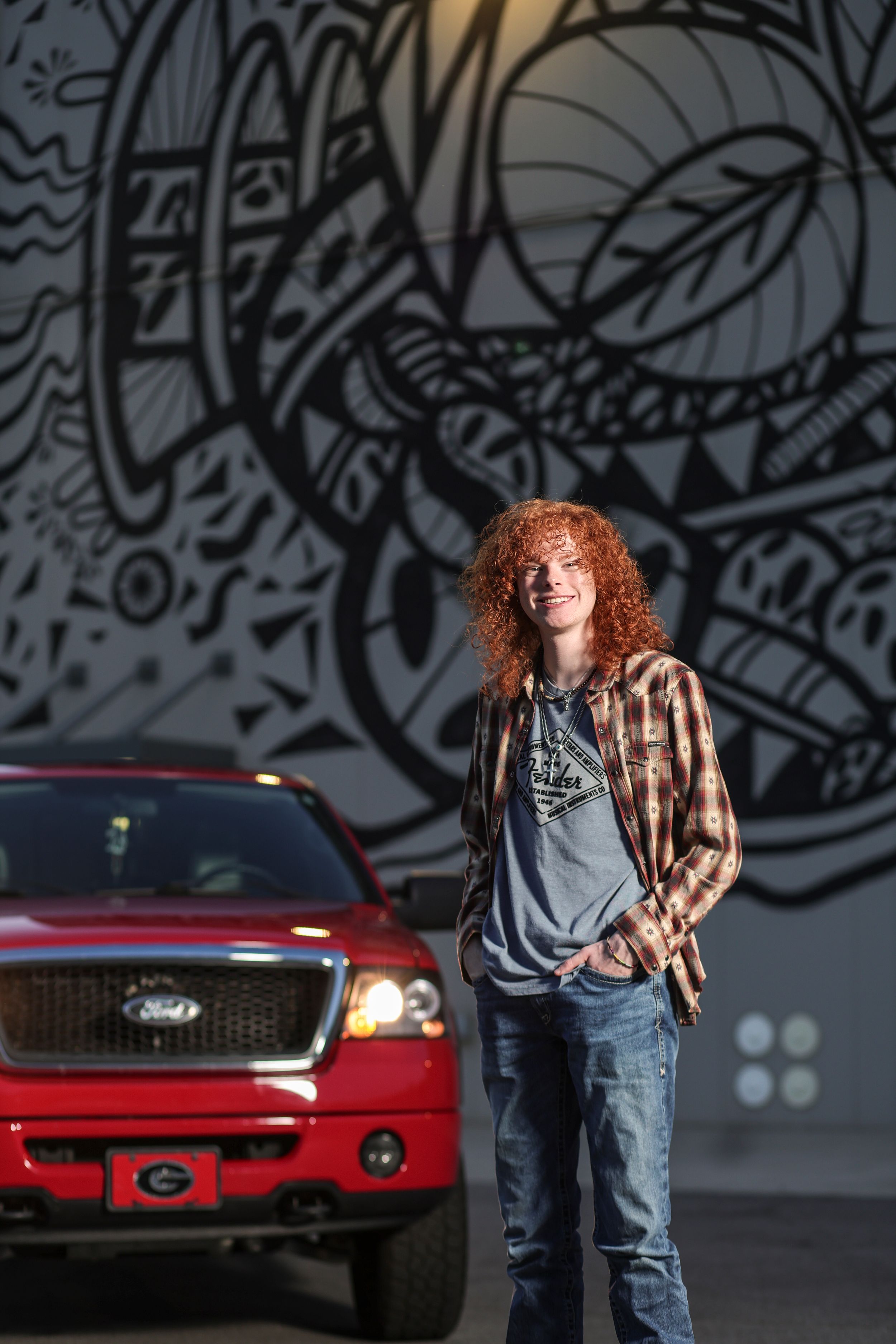 Person with red curly hair stands in front of large graffiti mural wall with red pickup truck parked nearby.