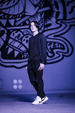 A person in all black clothing stands against a purple backdrop with intricate tribal patterns and designs.
