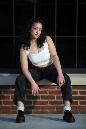 Person in white crop top and black jeans sits casually on brick ledge wearing platform shoes in artistic portrait.
