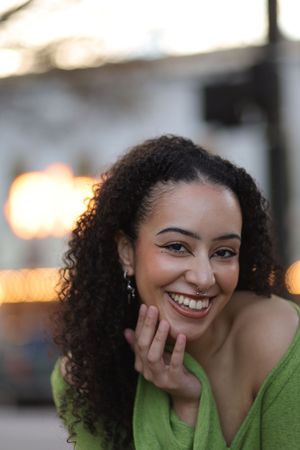 Person with natural curly hair wearing a green top smiles warmly while touching their face in warm lighting.