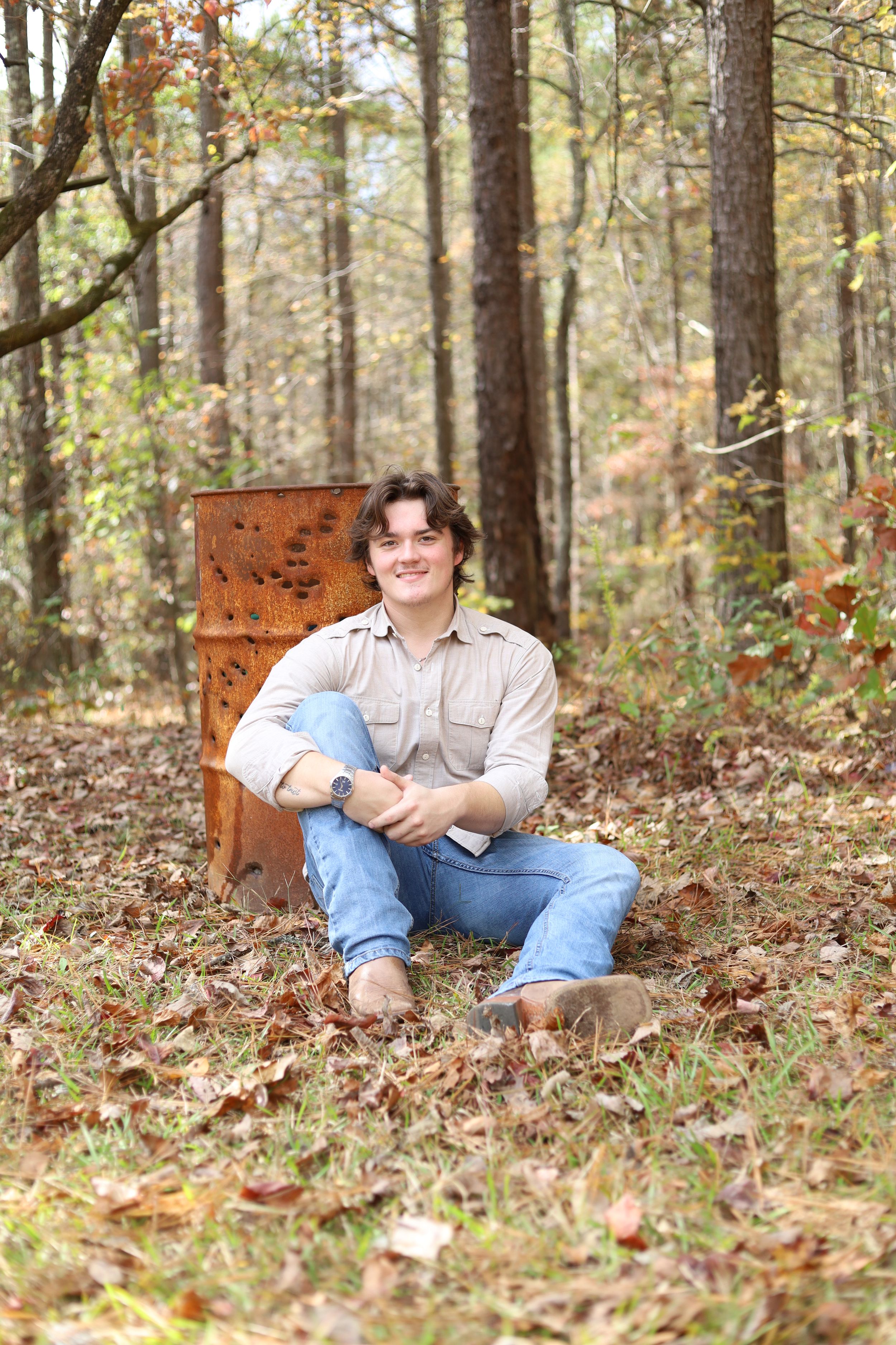 Someone sits casually against a rusty metal backdrop in an autumn forest setting surrounded by fallen leaves.