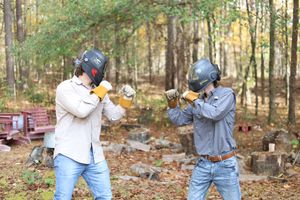 Two people in helmets and protective gear practice martial arts stances in a wooded outdoor setting.