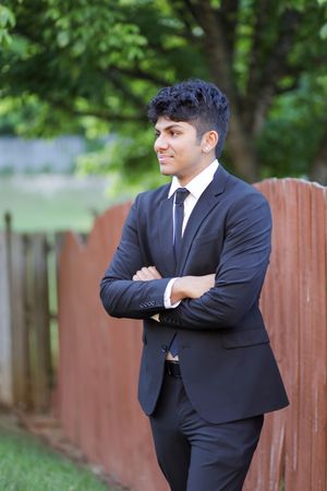 A young professional in a dark suit and tie standing outdoors with crossed arms and smiling beside a wooden fence.