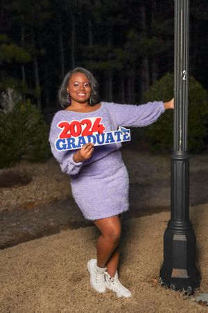 Someone poses in a lavender sweater dress with 2024 graduate text while leaning against a lamp post at night.