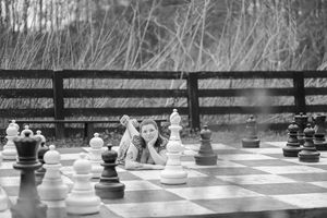 Giant outdoor chess game with white and black pieces on large board with wooden fence in background.