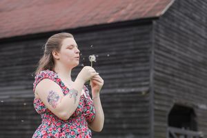 Person in floral dress blows dandelion seeds in front of rustic barn on cloudy day.