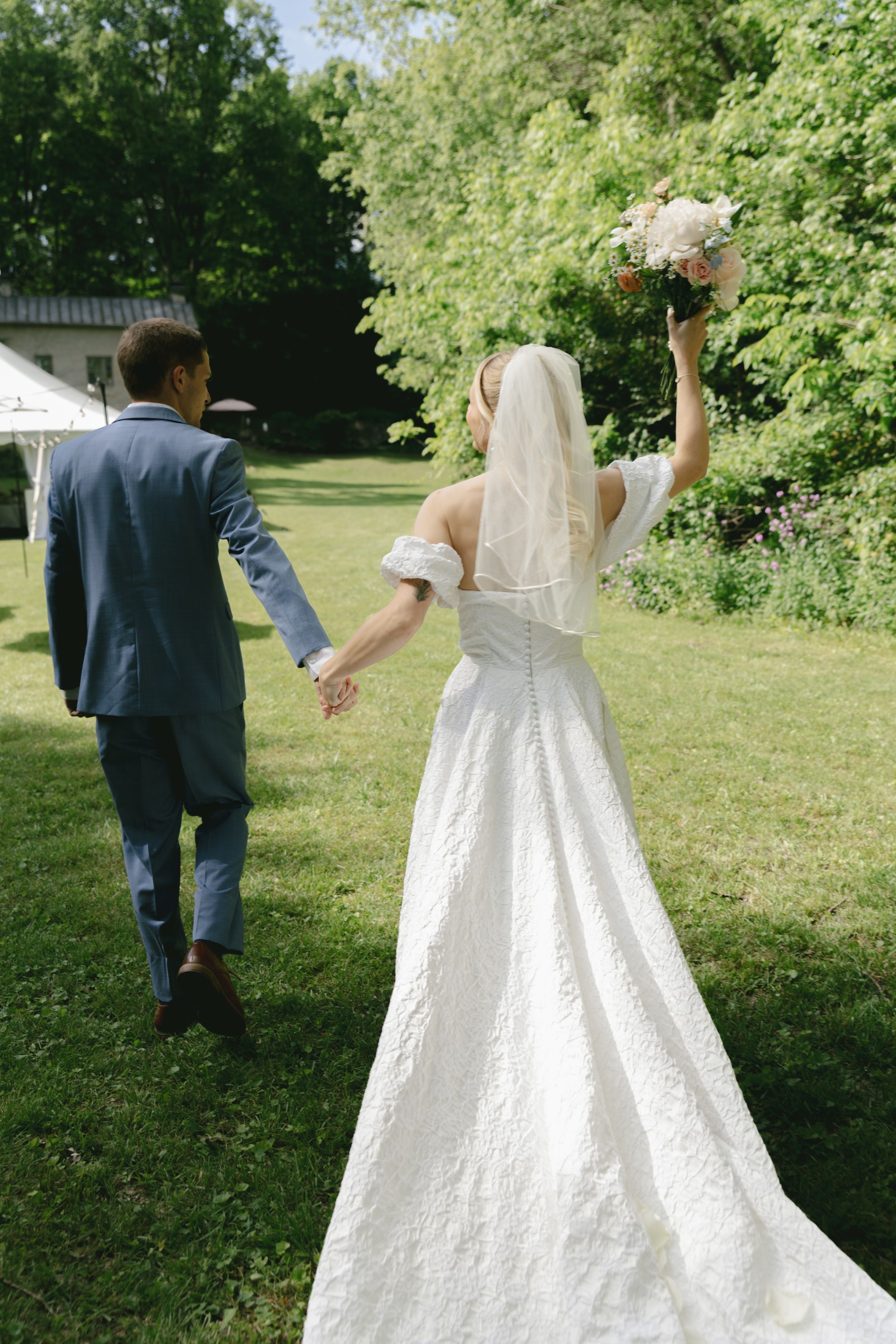 Newlyweds walk hand in hand across a lawn as bride holds bouquet aloft.