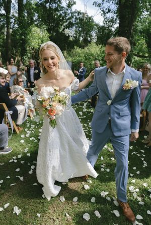 Newlyweds walk down the aisle amid flower petals while guests celebrate in an outdoor garden ceremony.