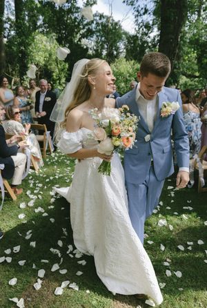 Wedding couple shares joyful laughter while walking through scattered petals in a sunlit garden setting.