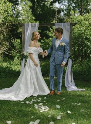 Bride in off-shoulder lace gown and groom in blue suit pose before white curtained arch outdoors.