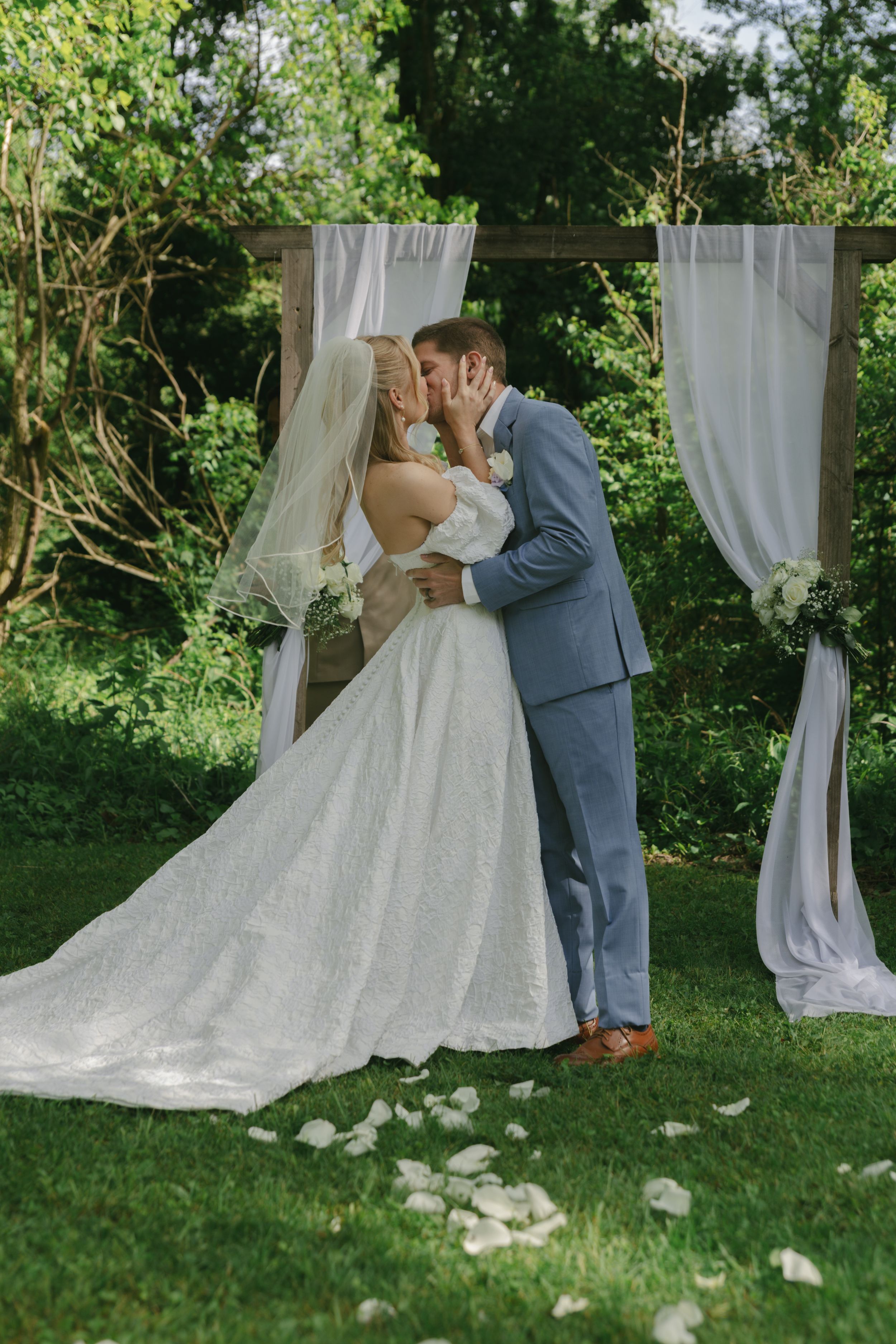 Romantic kiss between bride and groom under white draped arch with rose petals scattered below.