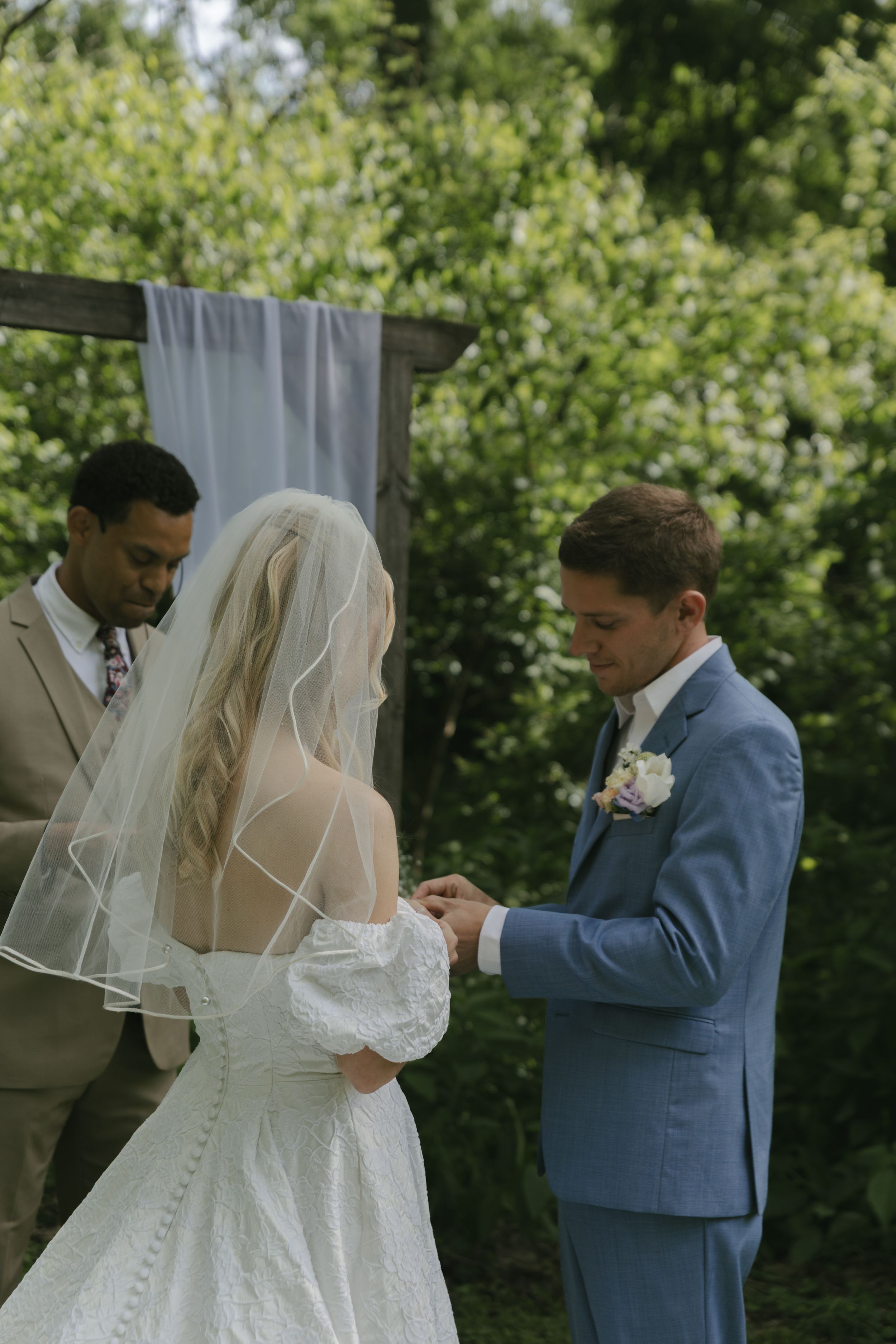 Wedding ceremony moment showing ring exchange with officiant in tan suit and white draped backdrop.