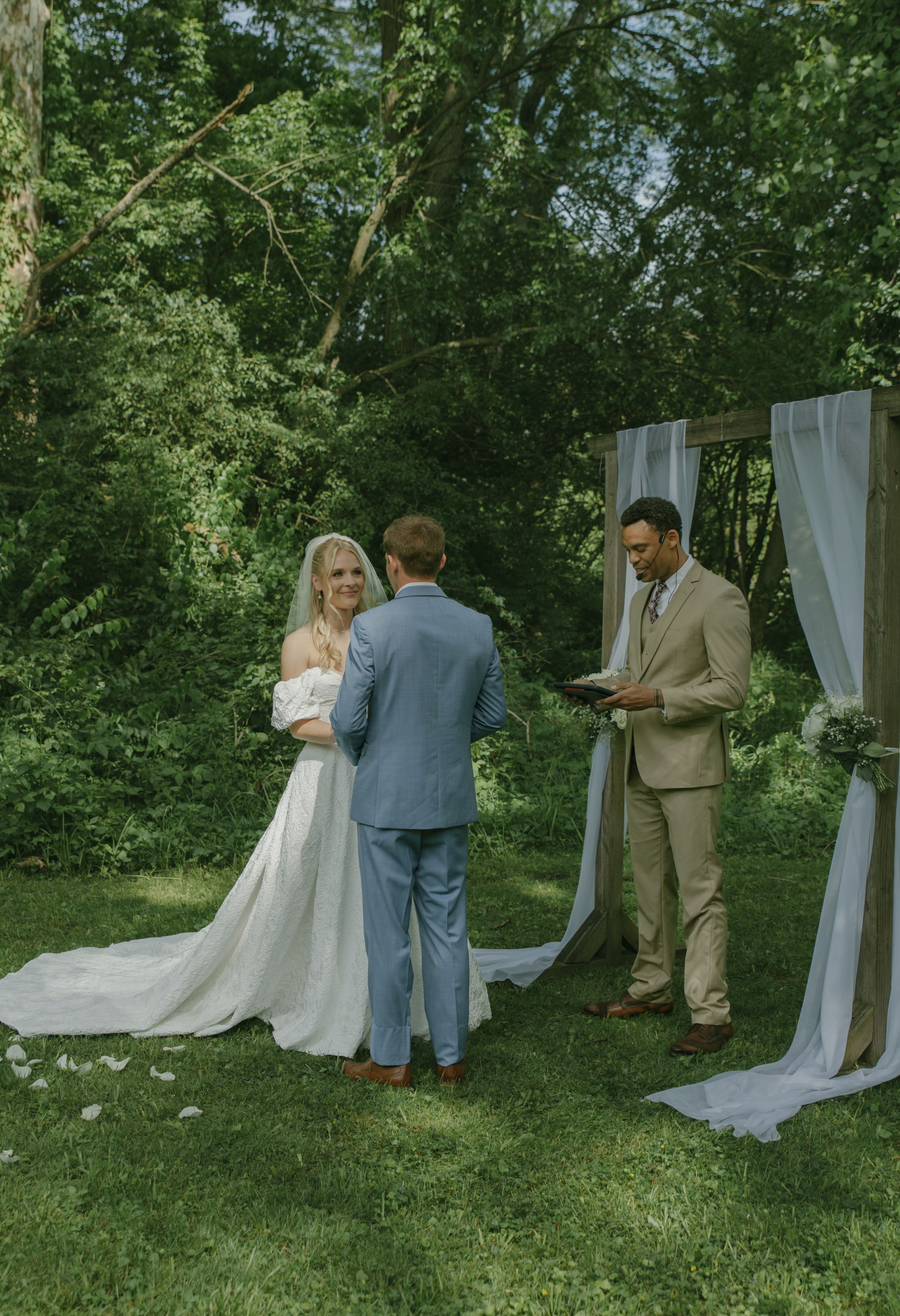 Outdoor wedding ceremony with officiant in tan suit and couple exchanging vows under white drapes.