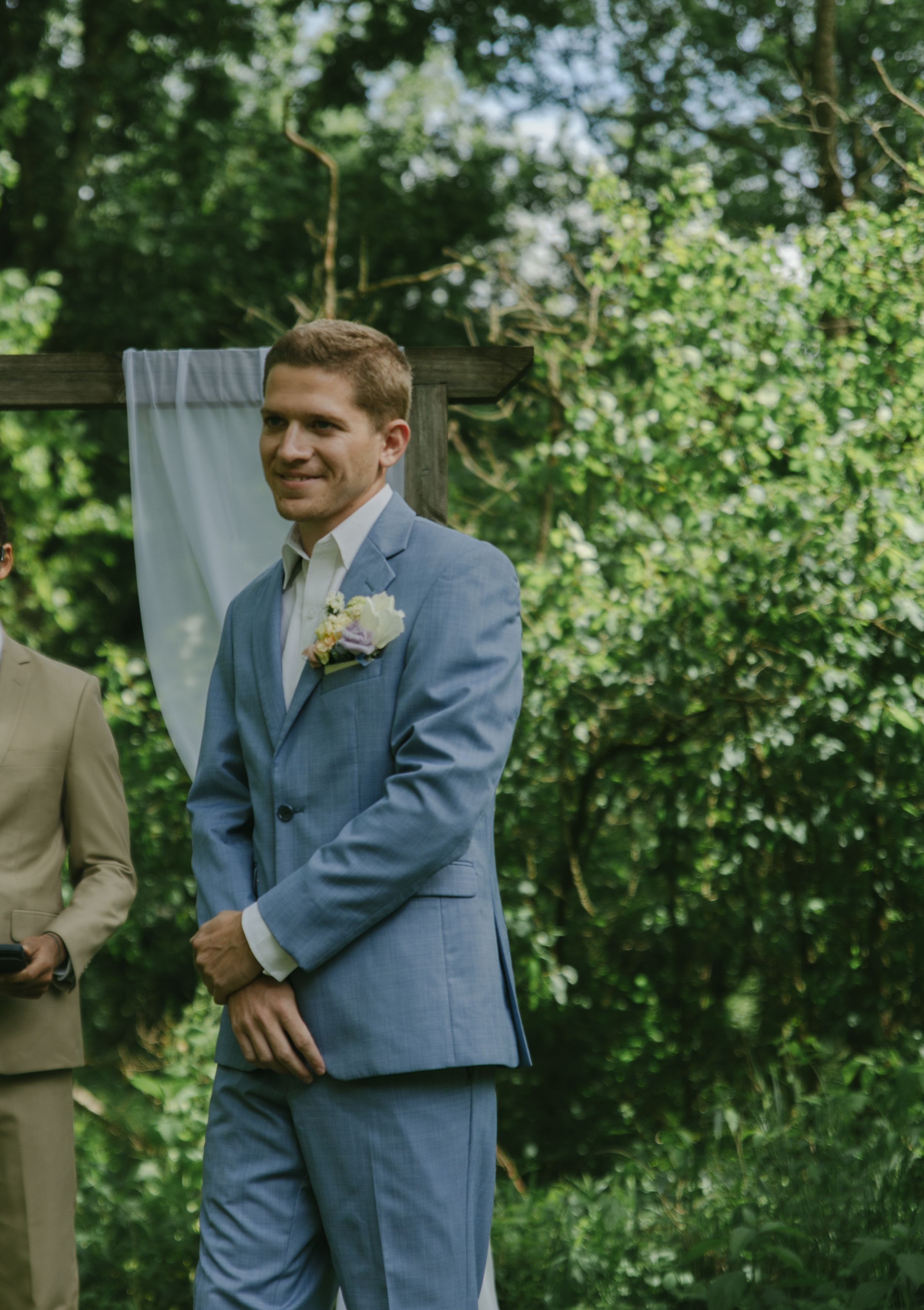 Groom in light blue suit stands at altar with boutonniere during outdoor ceremony.