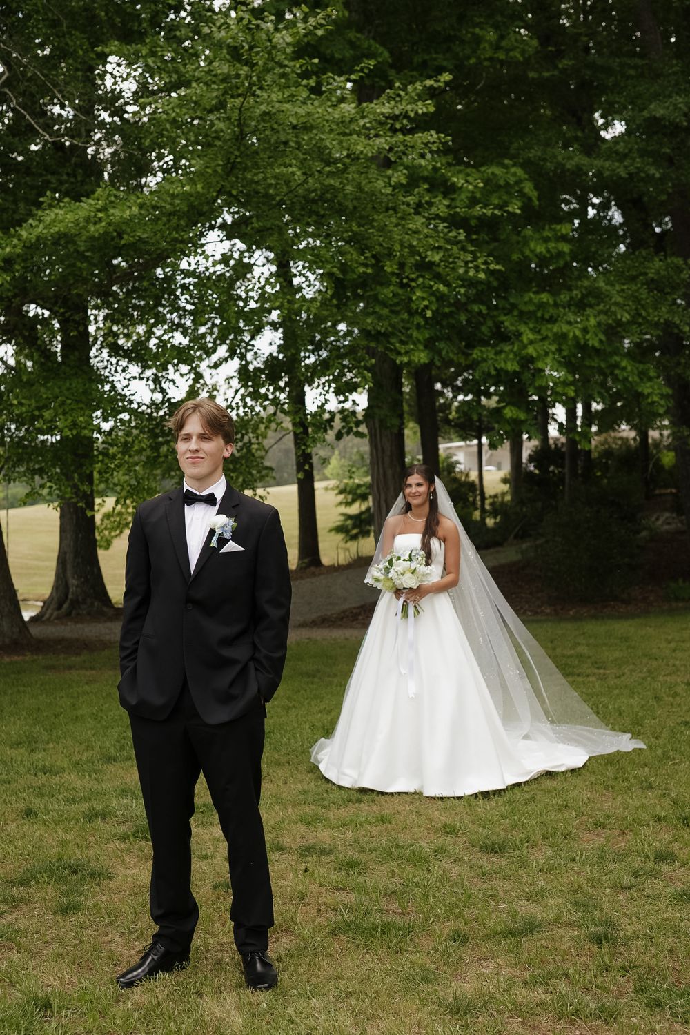 Bride in white gown with veil holds bouquet as groom in black tuxedo stands in foreground at outdoor wedding.