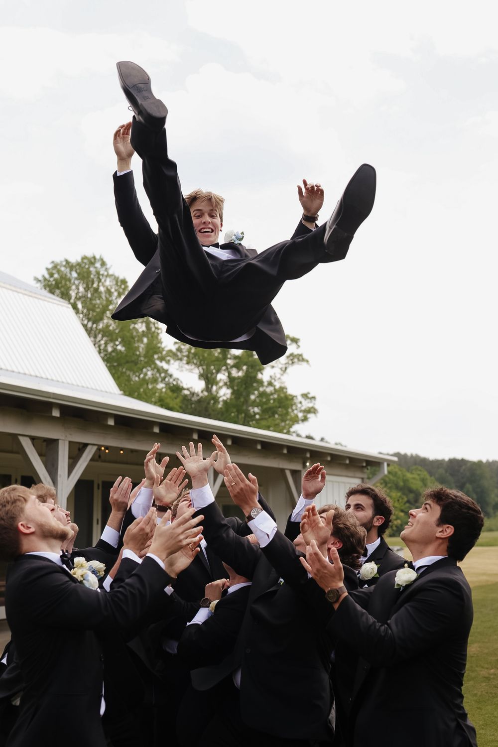 Groomsmen tossing groom into the air outdoors at a wedding celebration, all wearing black tuxedos.
