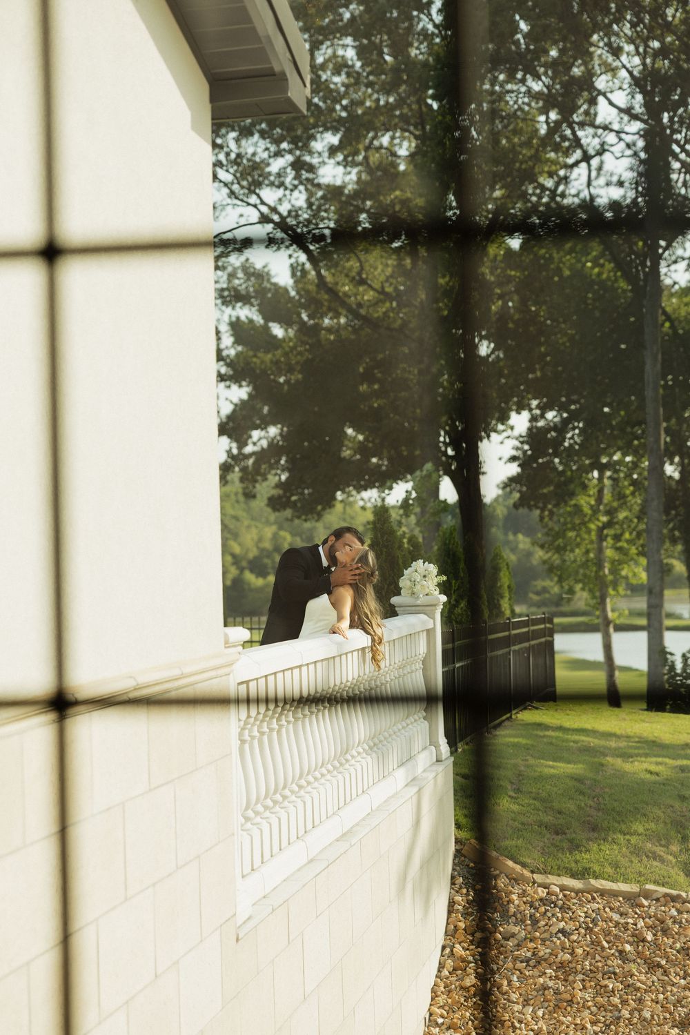 A couple sharing a romantic kiss on a white balcony, surrounded by lush green trees and manicured grounds.