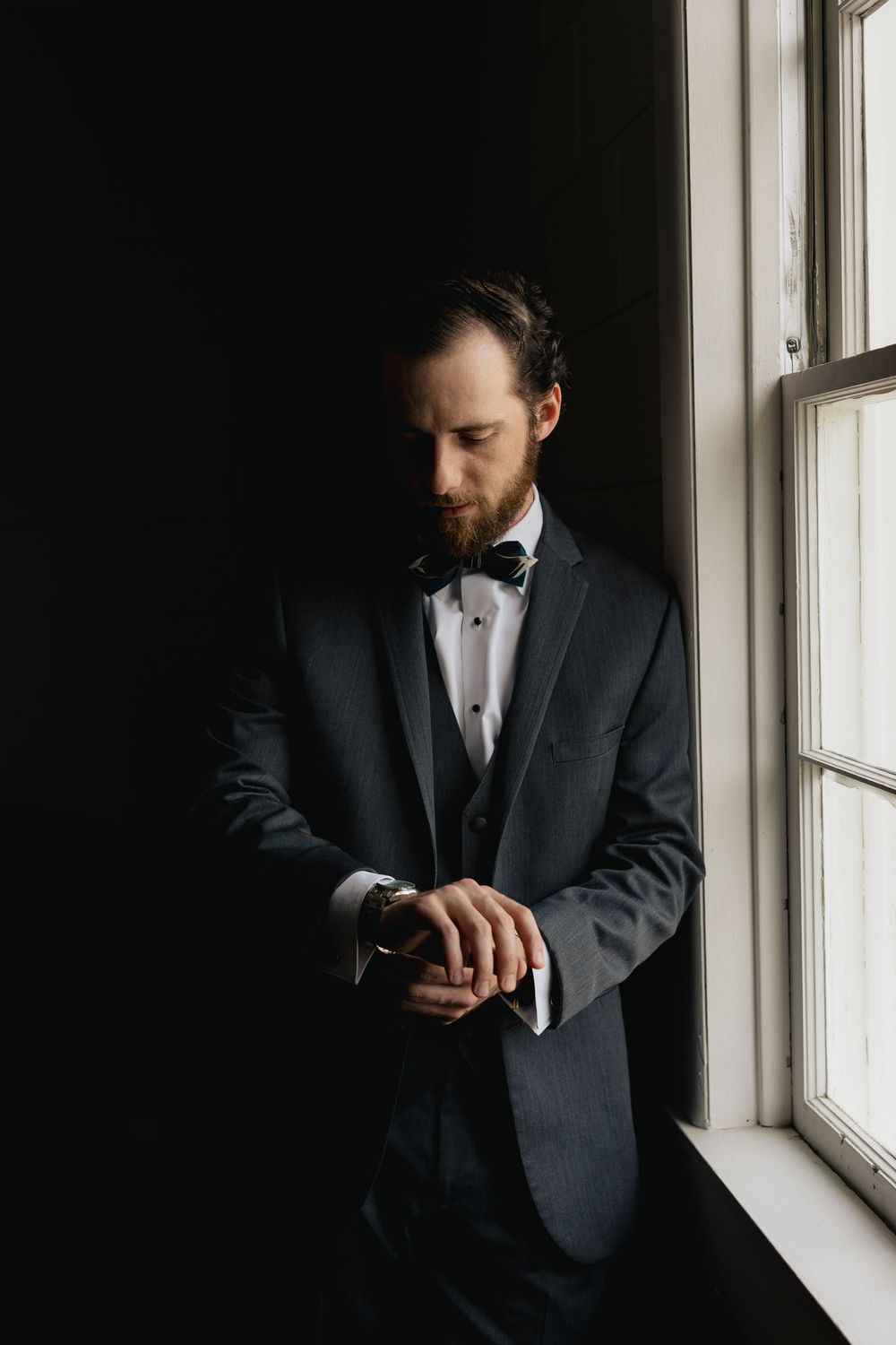 A dapper groom in a dark suit and bow tie checks his watch while standing by a window in moody lighting.