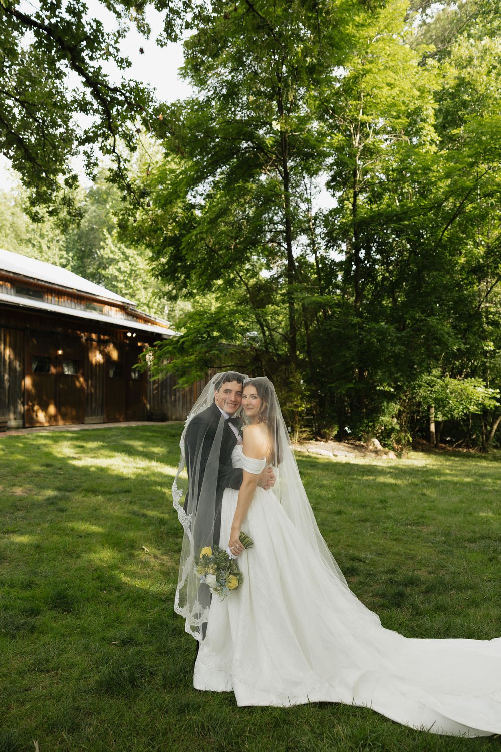 Bride and groom pose together outdoors on green lawn, bride wearing white gown with veil, holding bouquet near rustic barn.