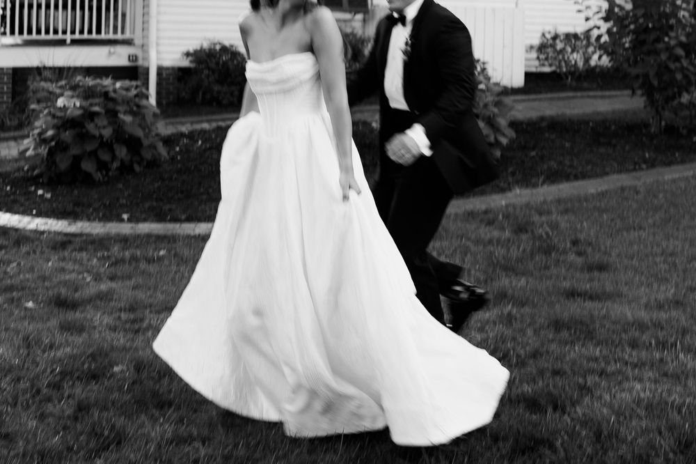 Black and white photo of a bride in a flowing white gown and groom in a tuxedo walking on a lawn.