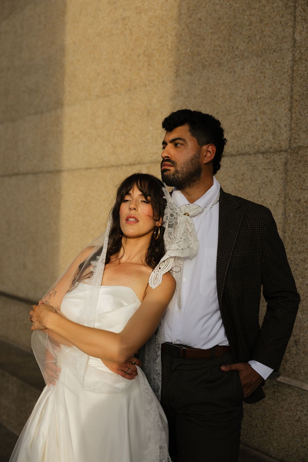 Bride in white gown and groom in dark suit pose elegantly against a sunlit stone wall.