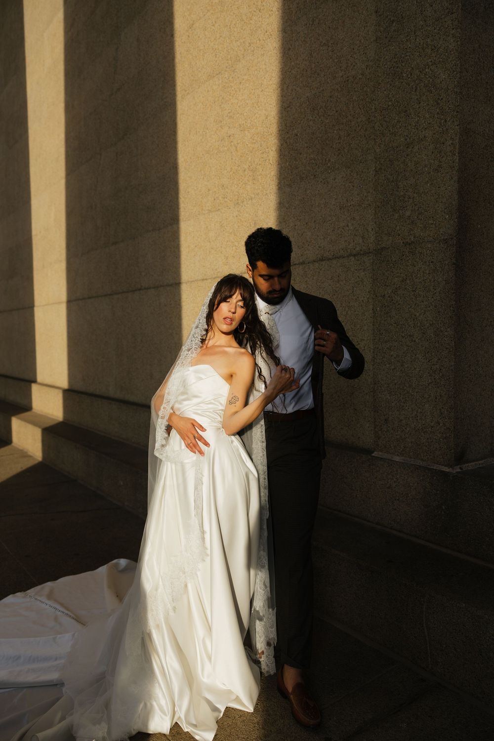 A bride in a white gown and veil embraces her groom in a dark suit, bathed in golden sunlight against stone columns.
