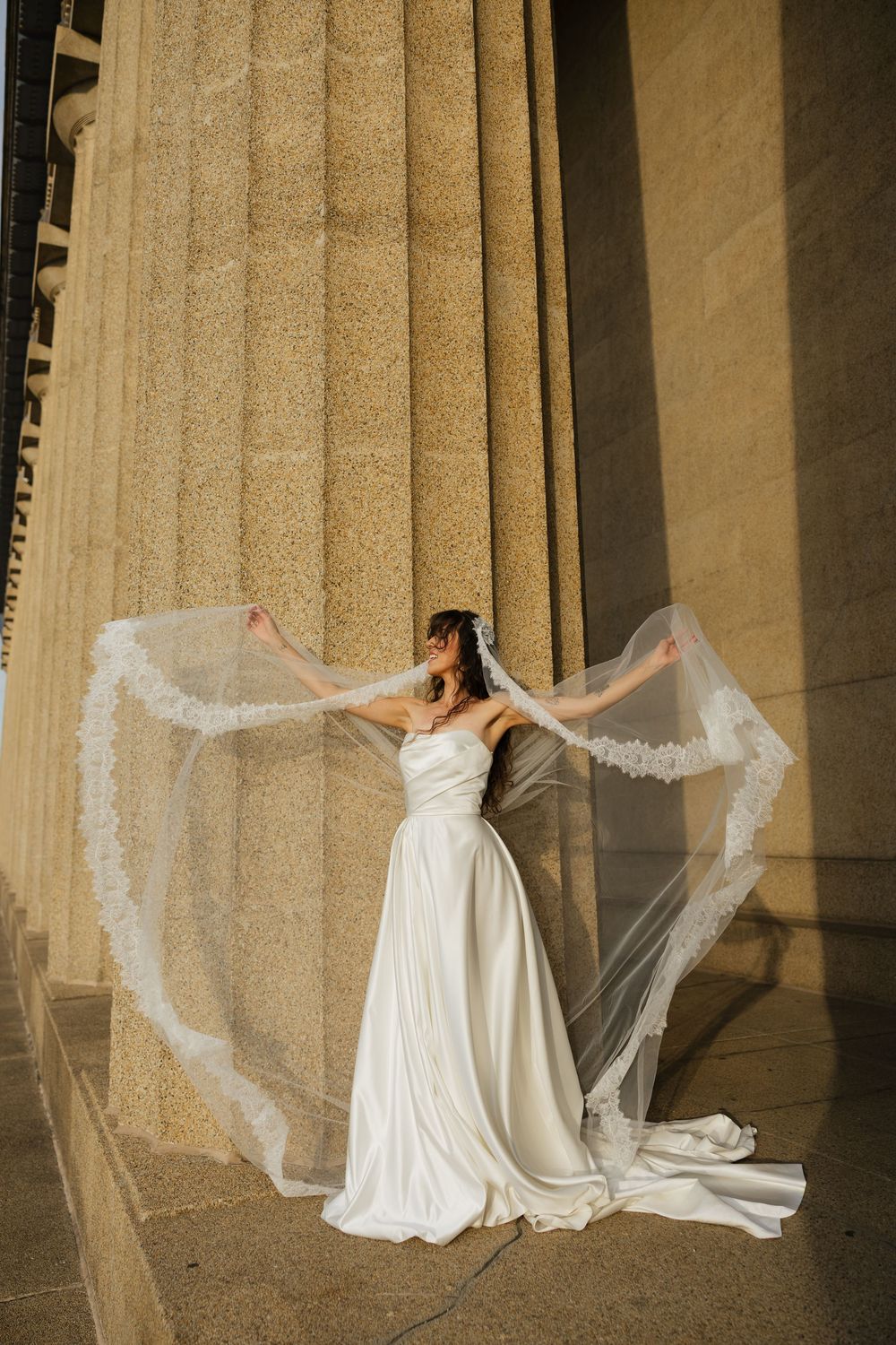 Bride in white strapless gown holds flowing lace veil wide beside grand stone columns in golden light.