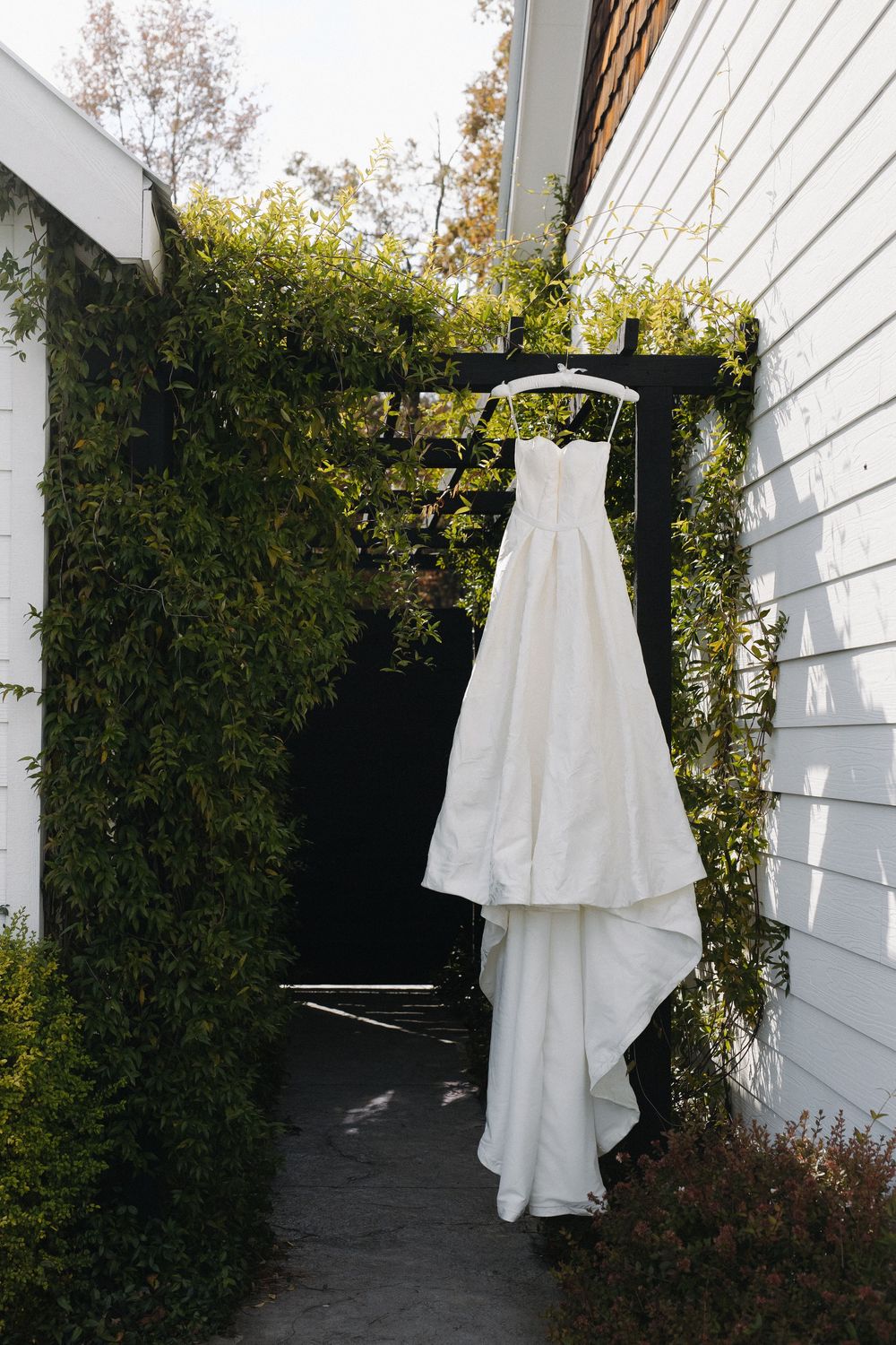 White wedding dress hanging outdoors against white siding and green foliage.
