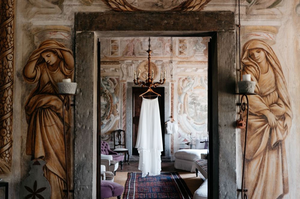 A white wedding dress hangs in an ornate doorway framed by classical carved angel figures and baroque wall paintings.