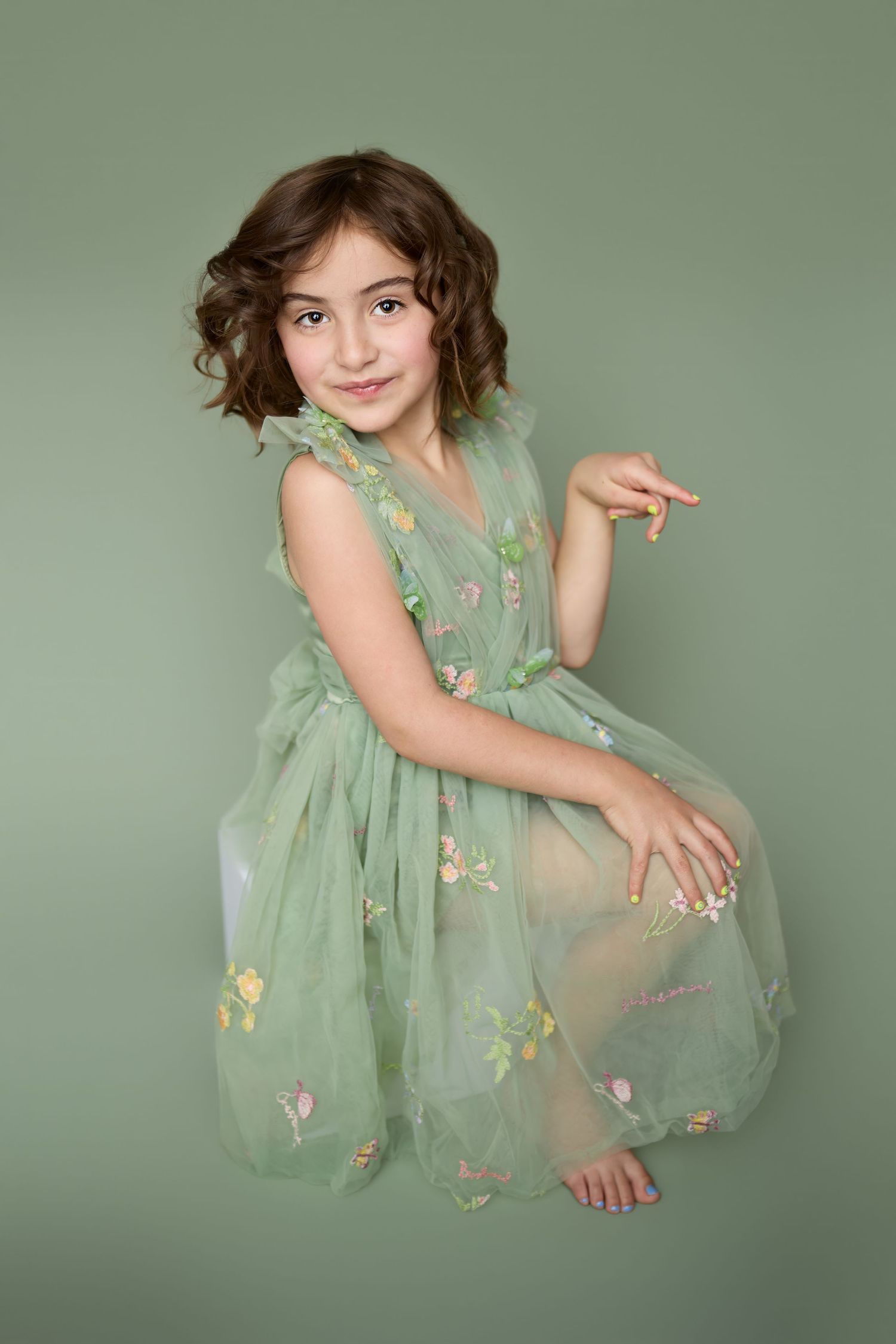 Young child wearing a flowy mint green tulle dress with floral embellishments poses against sage green background.