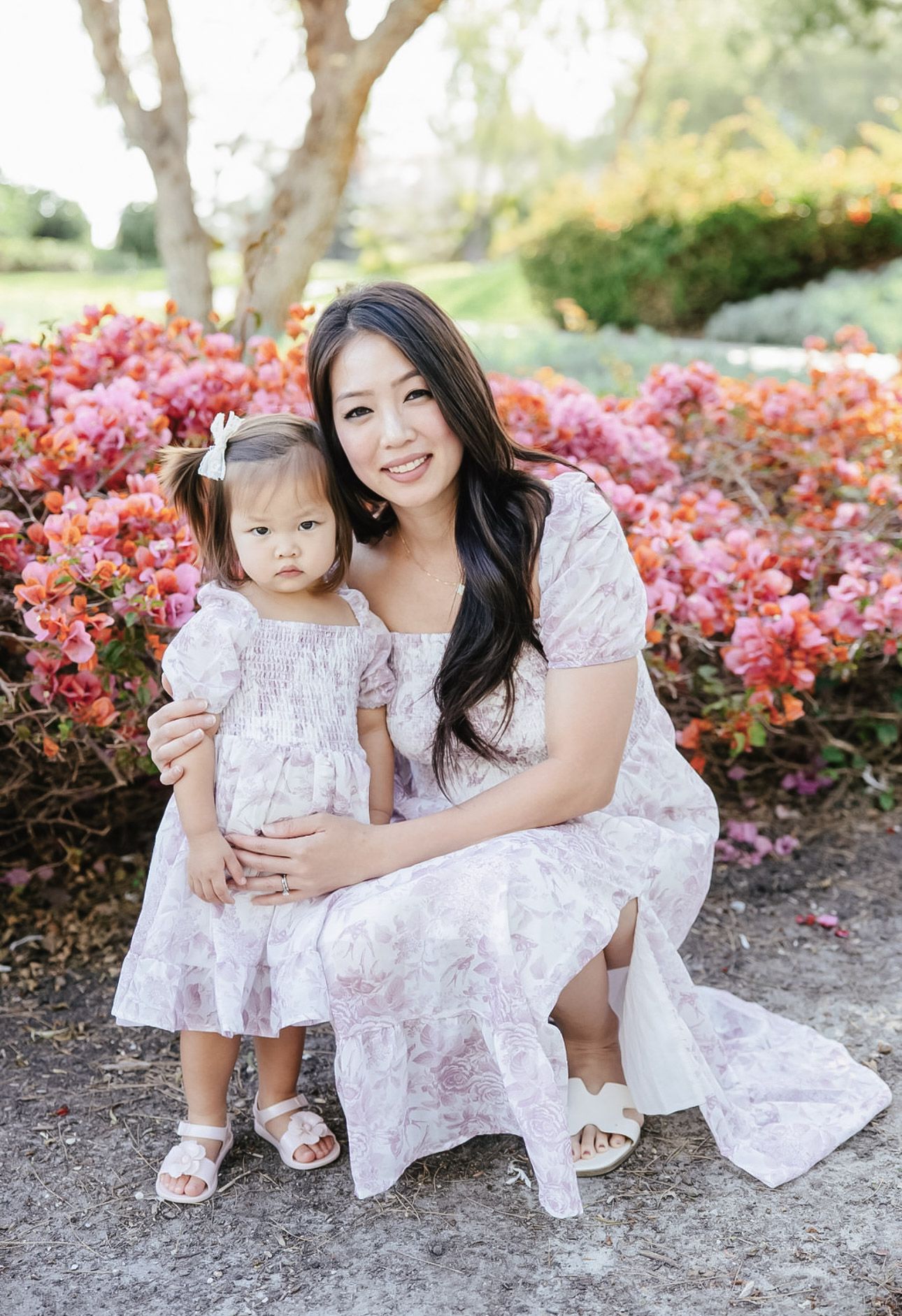 Mother and daughter wearing matching white floral dresses pose together among pink flowering bushes in a garden setting.