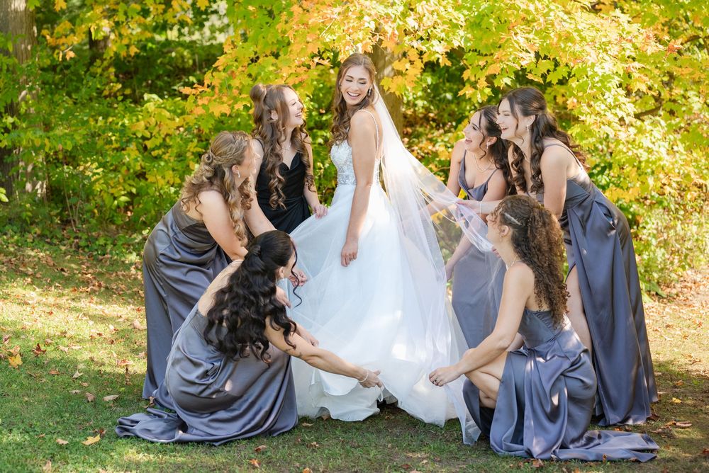 Bridesmaids in grey dresses help arrange the bride's gown outdoors during a fall photoshoot.