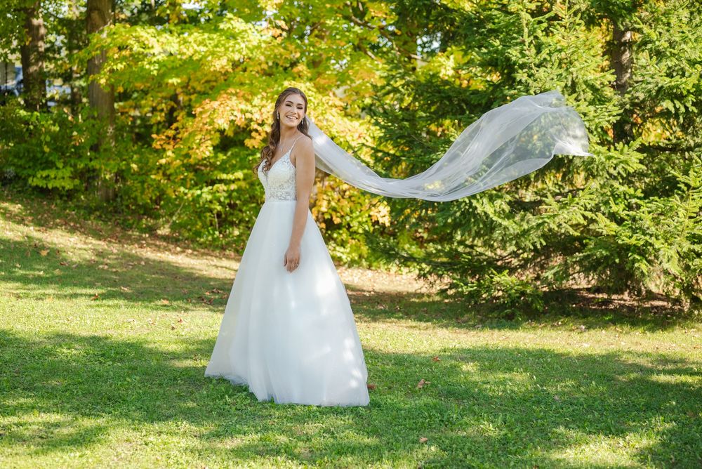 A bride in a flowing white wedding dress spins gracefully on a sunny lawn with autumn trees in the background.