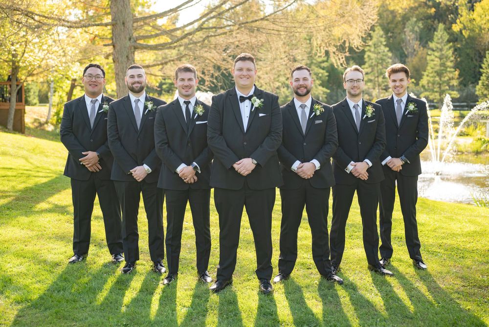 A group of seven groomsmen in black suits and bowties stand in a line on a sunny autumn lawn.