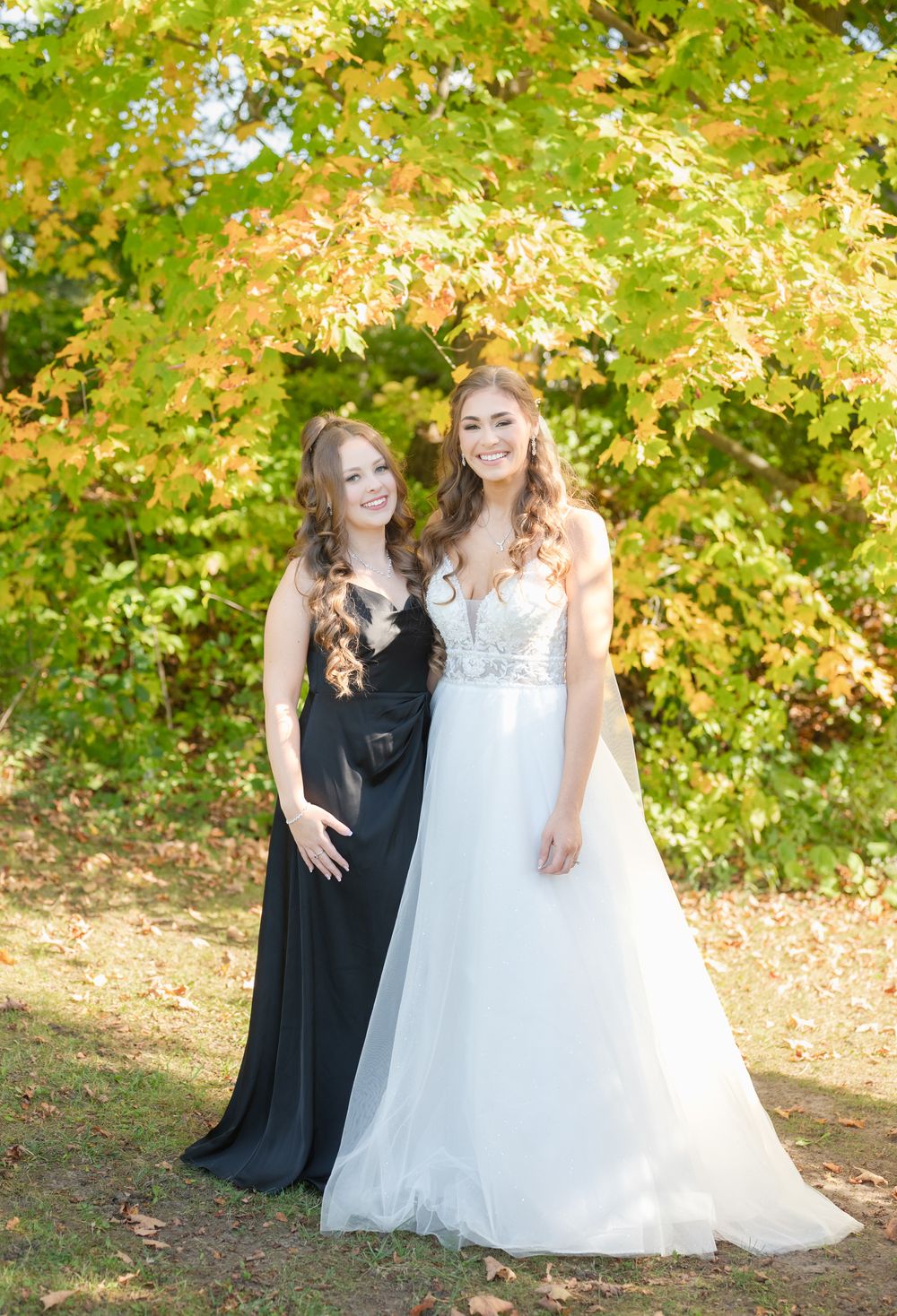A bride in a white gown and bridesmaid in black standing together among autumn foliage.