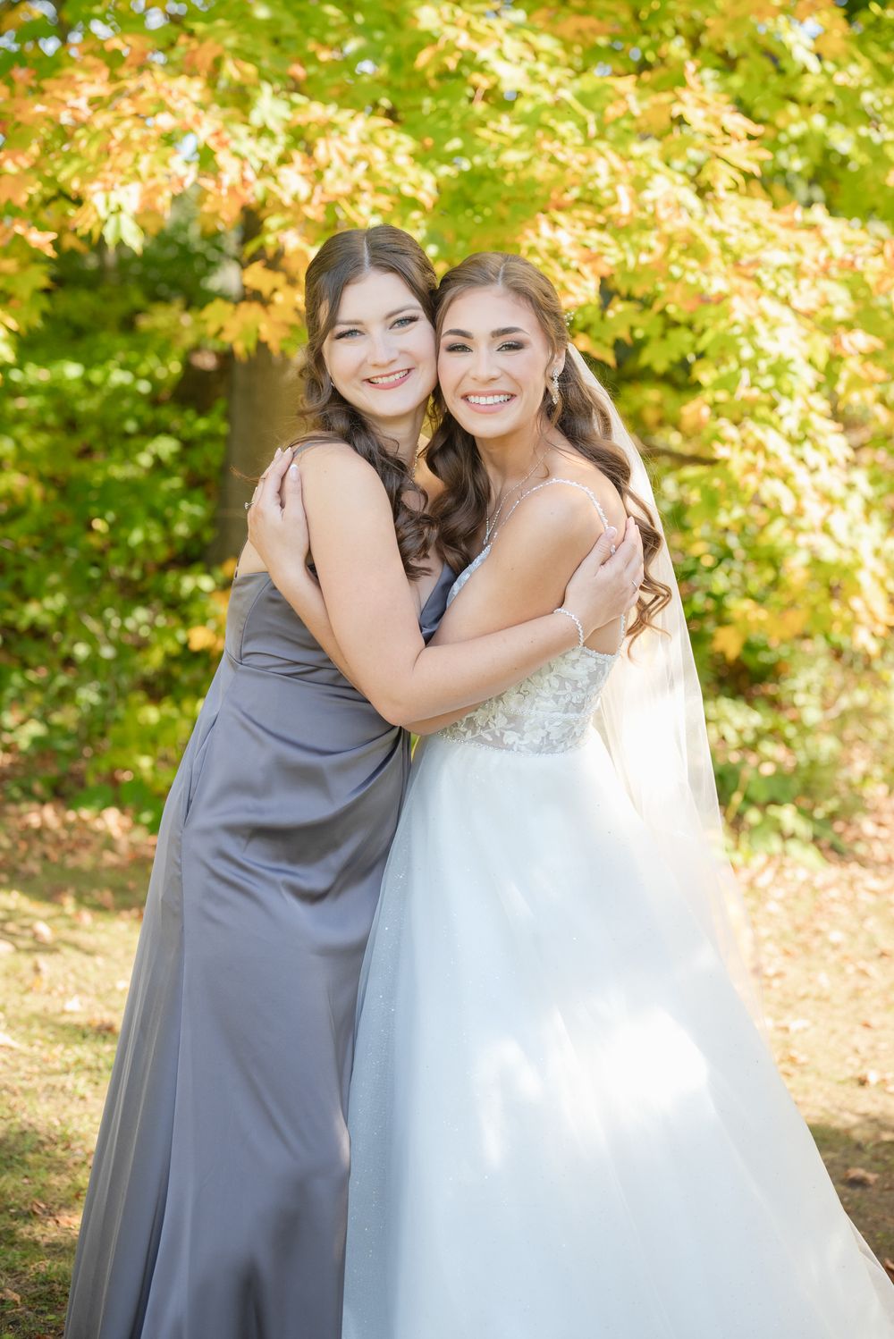 A bride in white gown embraces a bridesmaid in grey dress against autumn foliage background.