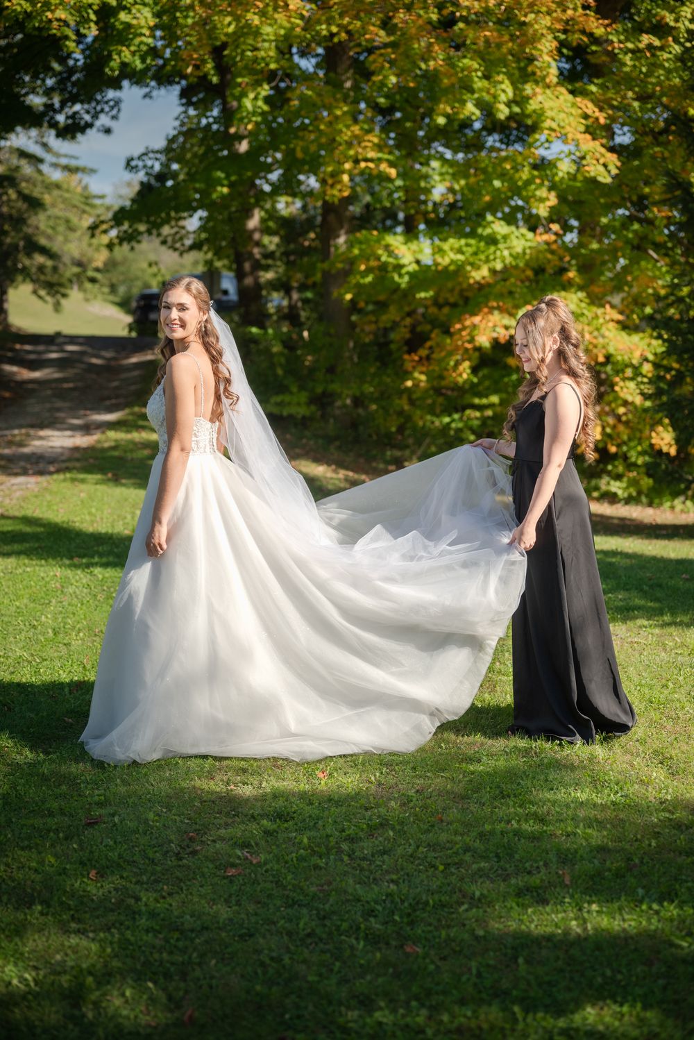 Bride in flowing white gown poses with bridesmaid against autumn foliage.