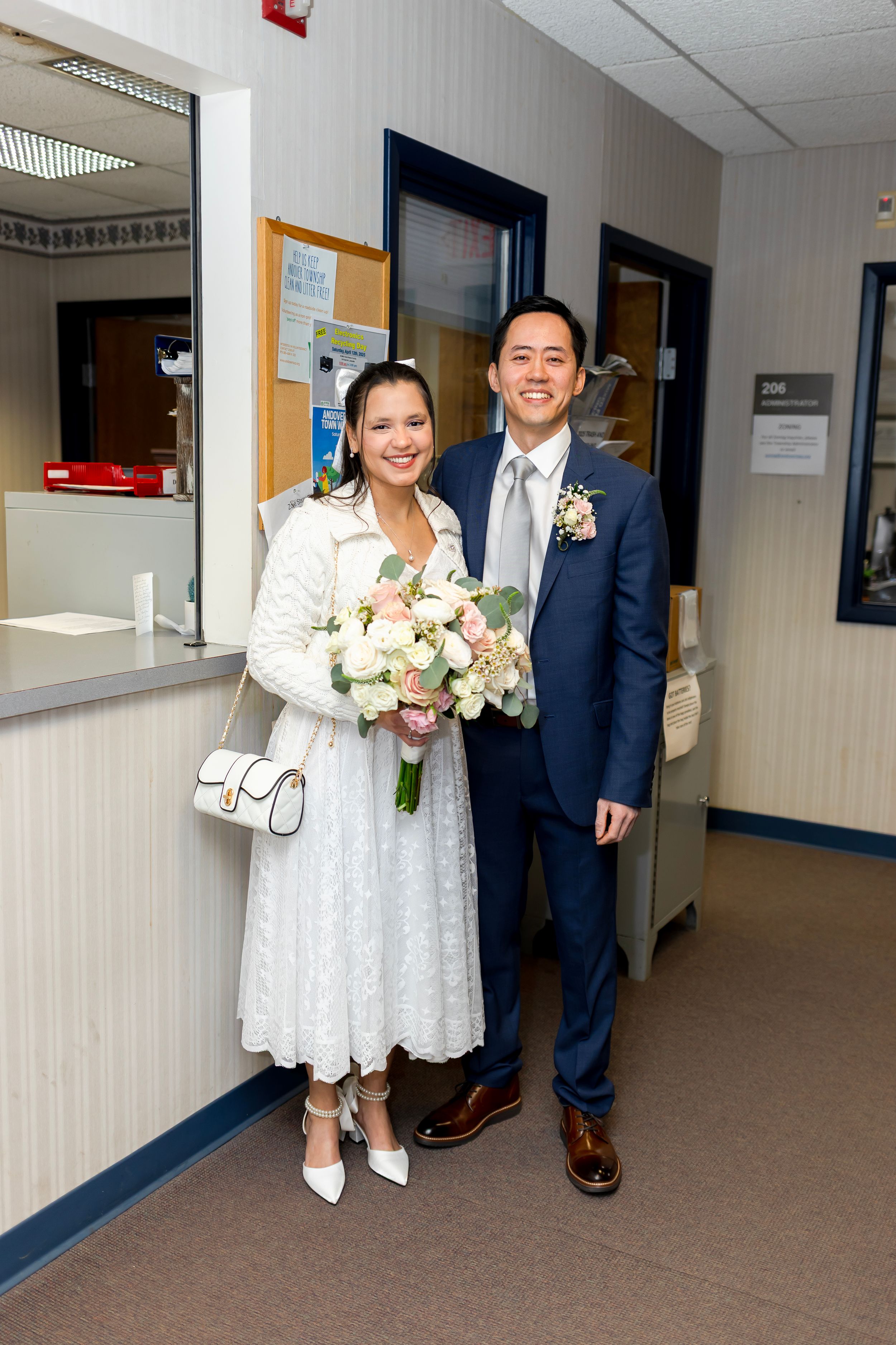 A couple in wedding attire stands in a hallway - the bride in a white lace dress holding flowers, groom in a navy suit.