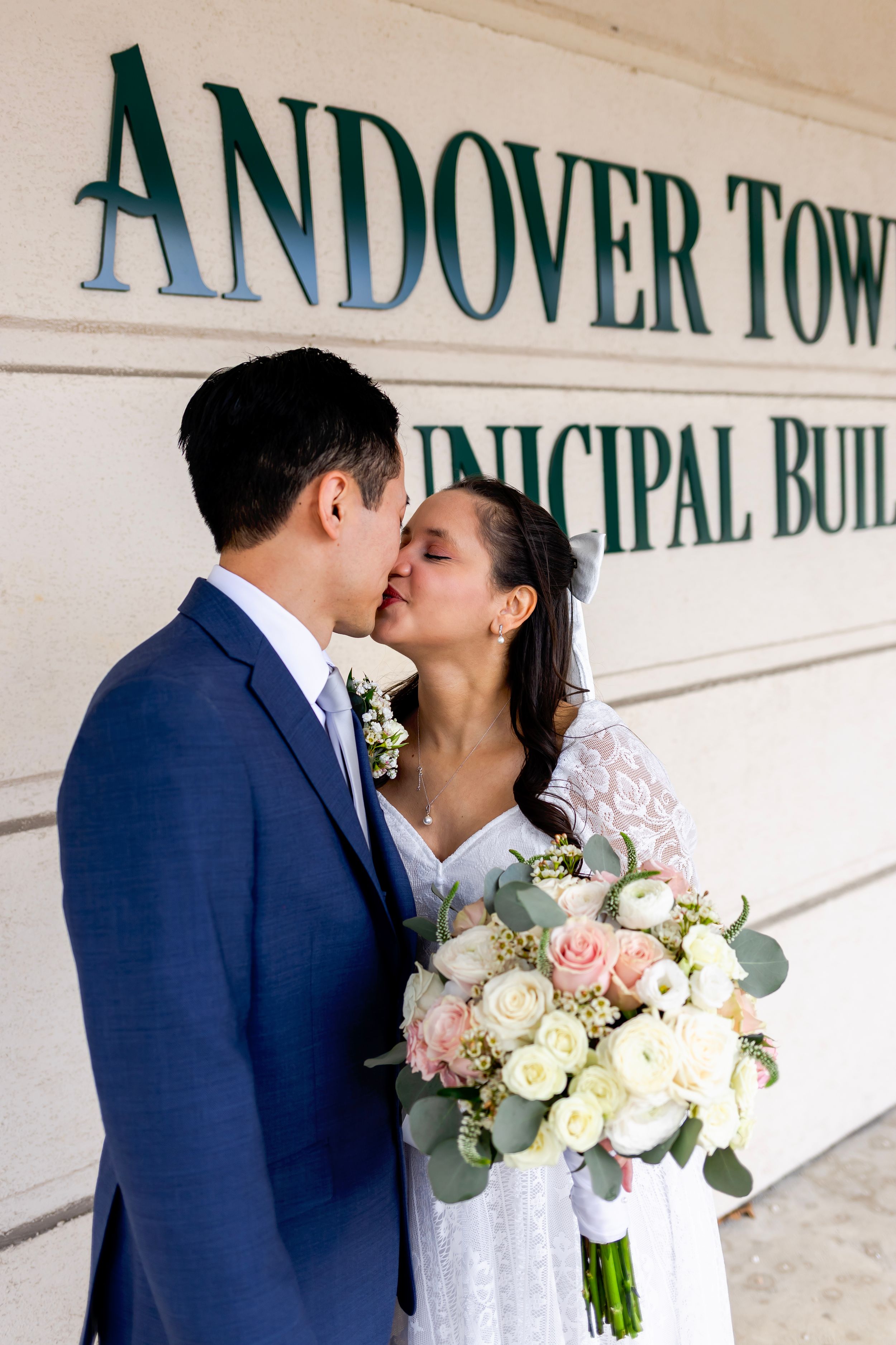 A romantic wedding couple kisses outside Andover Town Municipal Building with bride holding a peach and white bouquet.