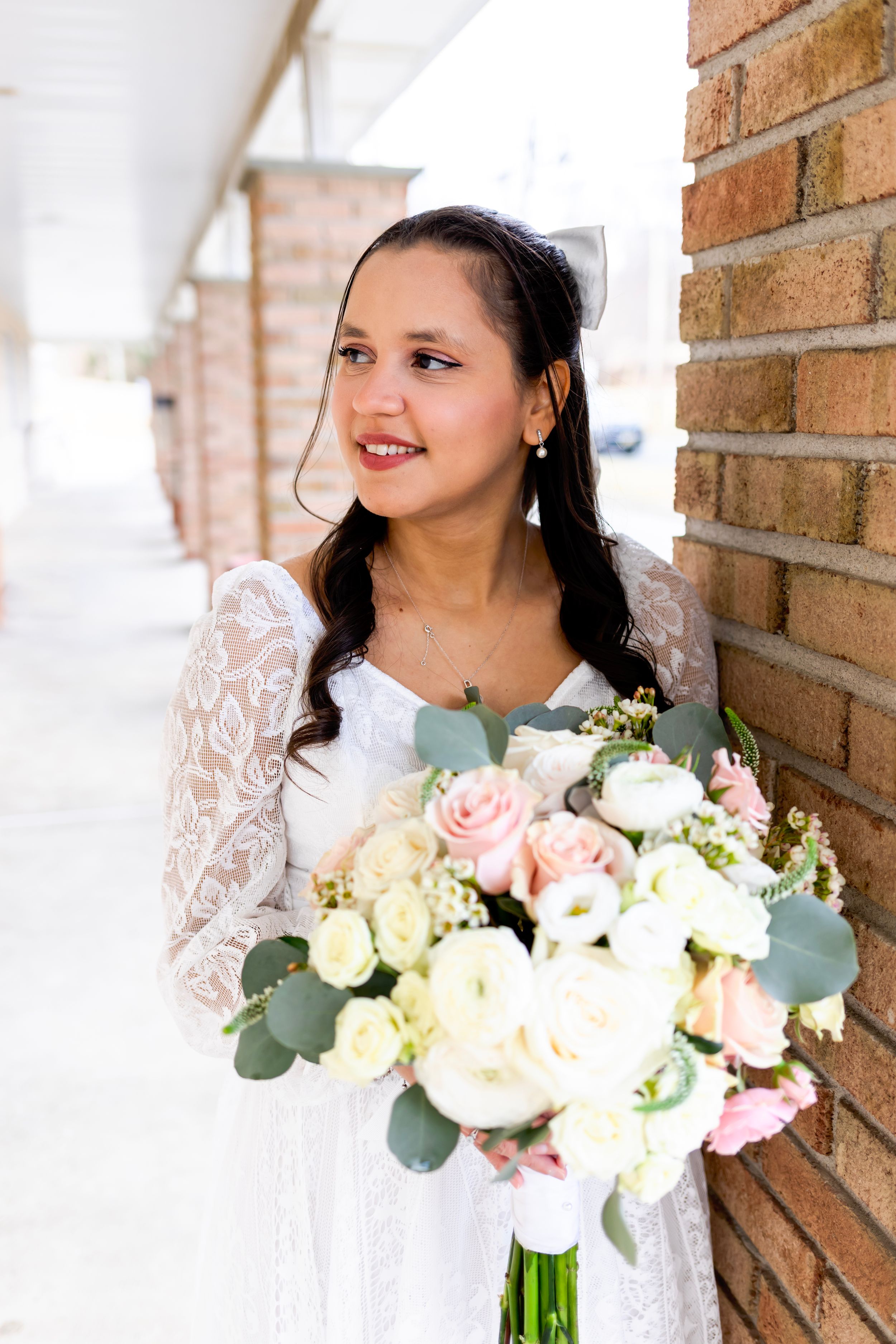 A bride in a white lace wedding dress holds a beautiful bouquet of blush pink roses and eucalyptus against a brick wall.