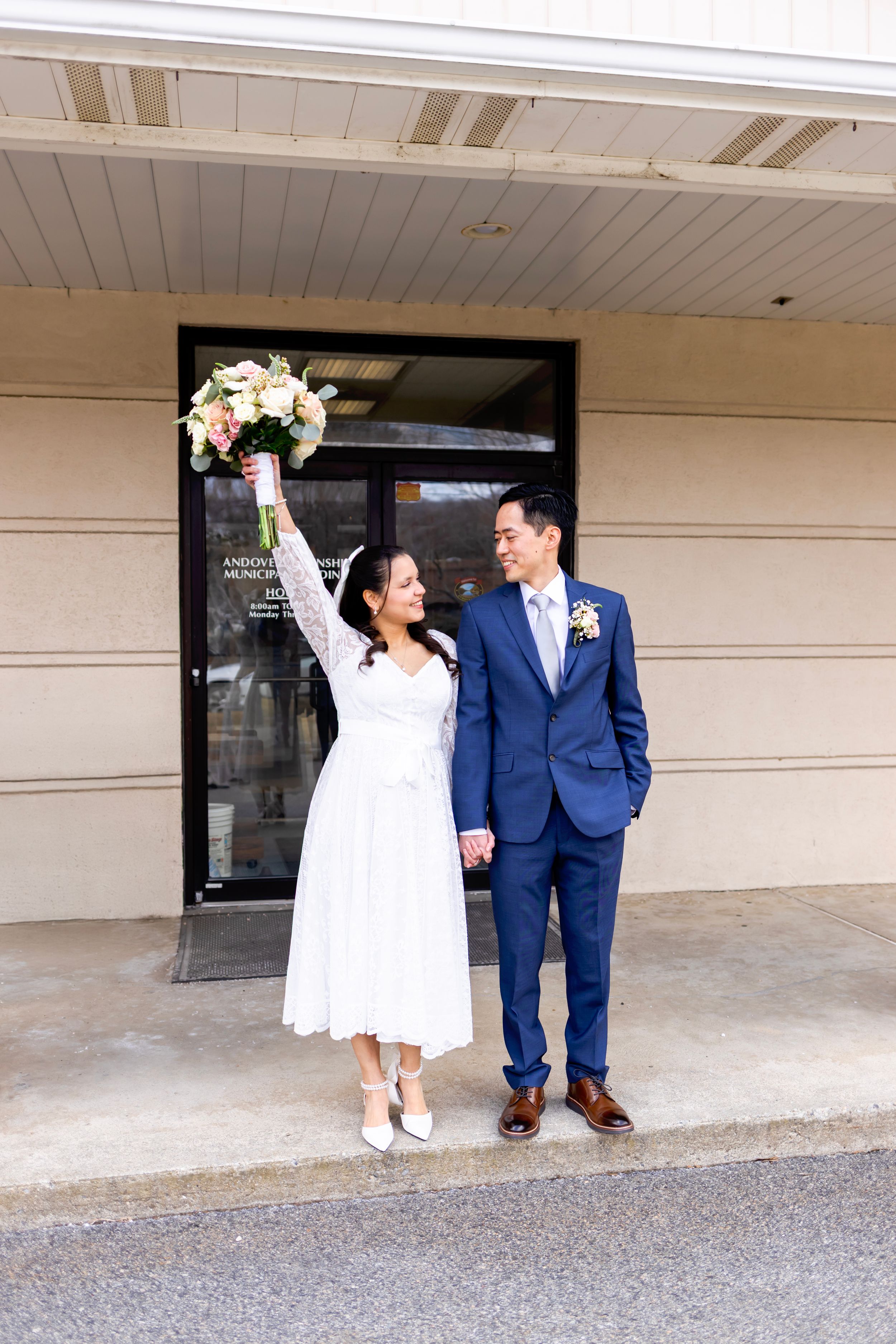 A couple celebrates their wedding day outside a building, with the bride in white holding up her bouquet and groom in navy suit.