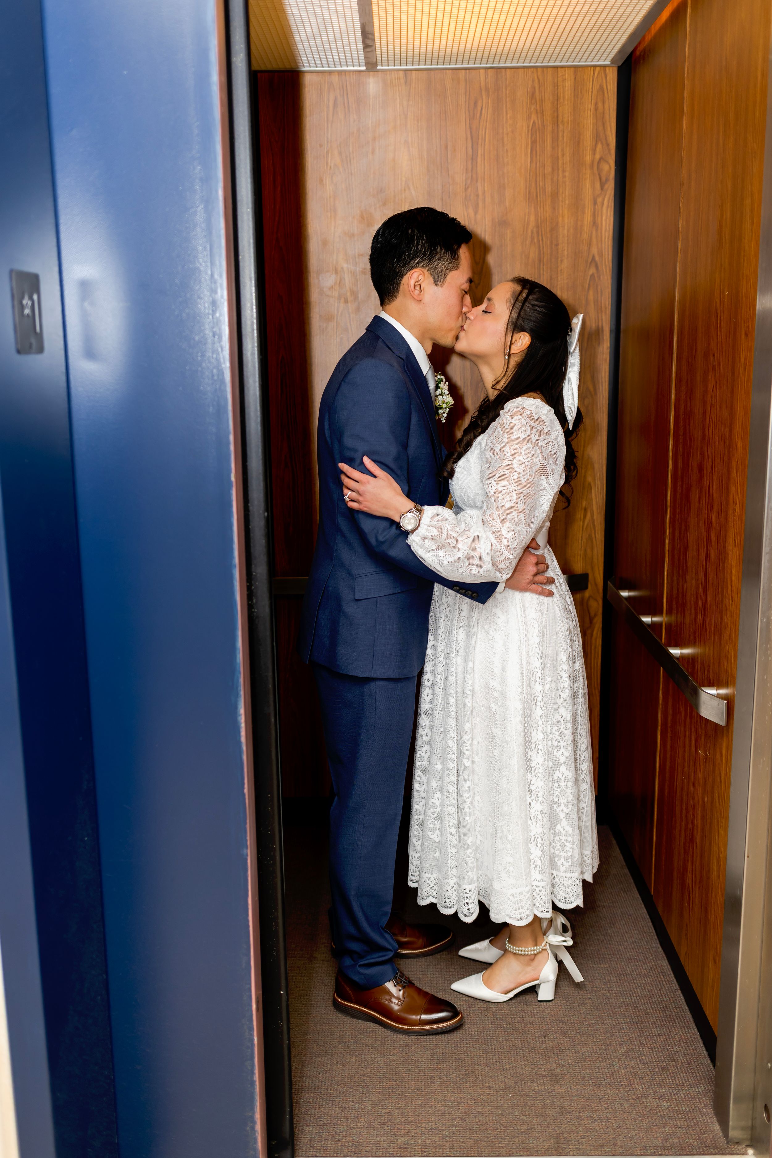 A couple shares an intimate wedding kiss in a hallway, the bride wearing a white lace dress and groom in a navy suit.