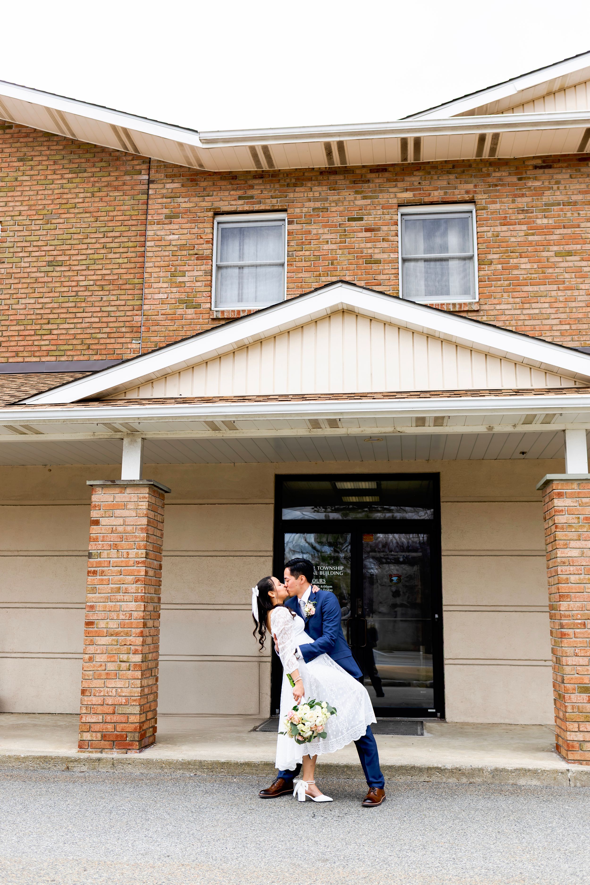 A romantic embrace outside a brick building with white trim and two windows, featuring a bouquet of white flowers.