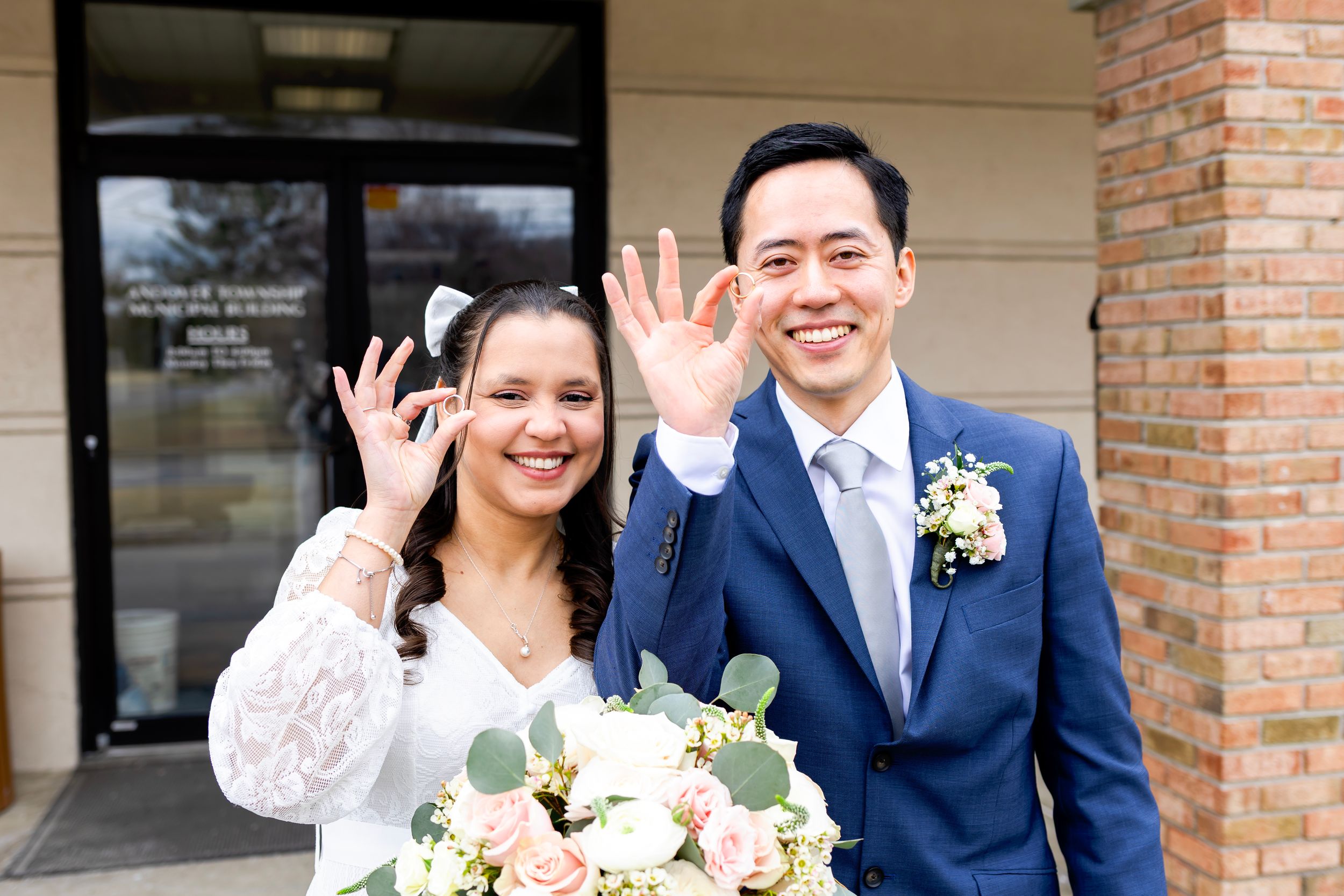Happy couple waves at camera while holding pink and white wedding bouquet in front of brick building.