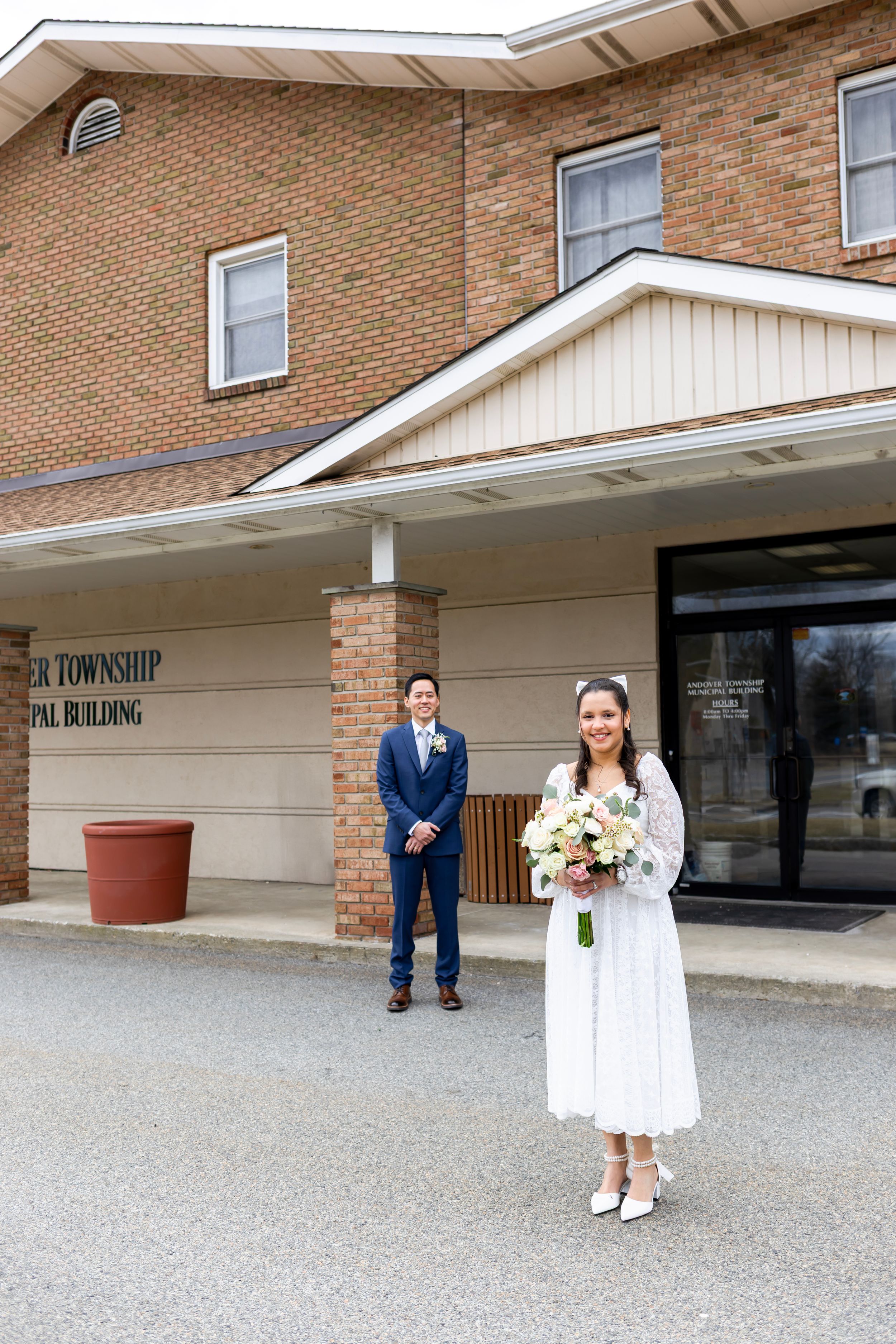 Two people stand outside a brick township municipal building, wearing formal attire and holding a bouquet.