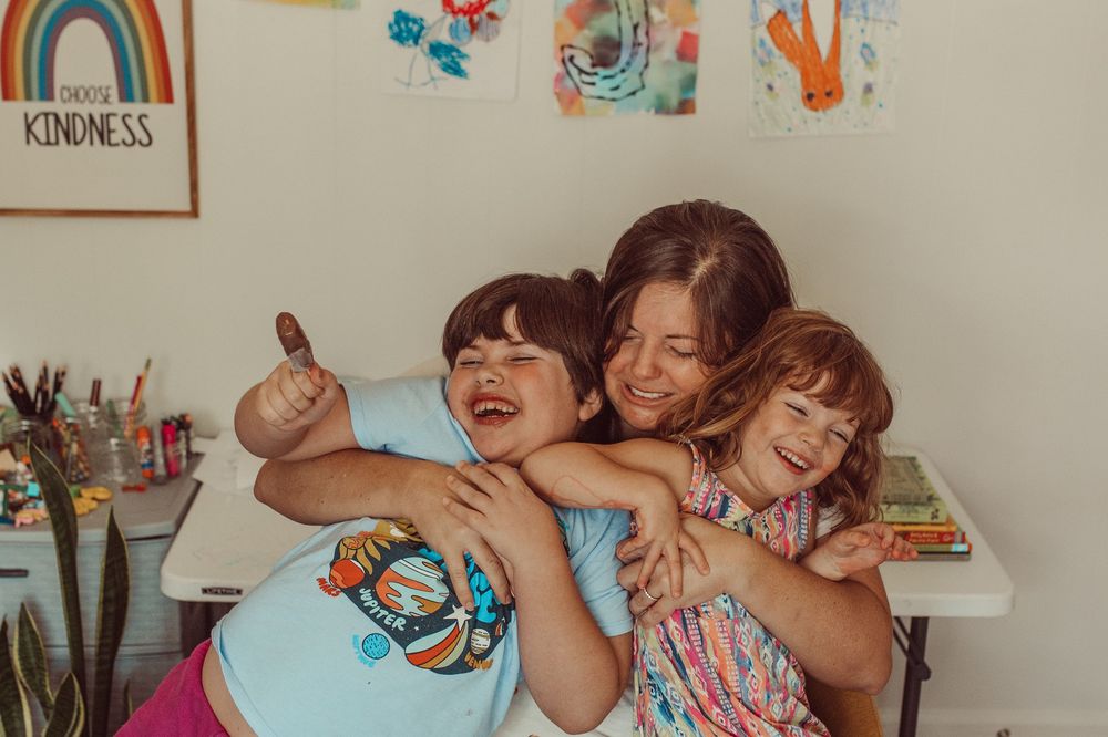 Mom and children share a joyful moment of laughter and hugs while playing together in a colorful room with wall art. WHITNEY POPP