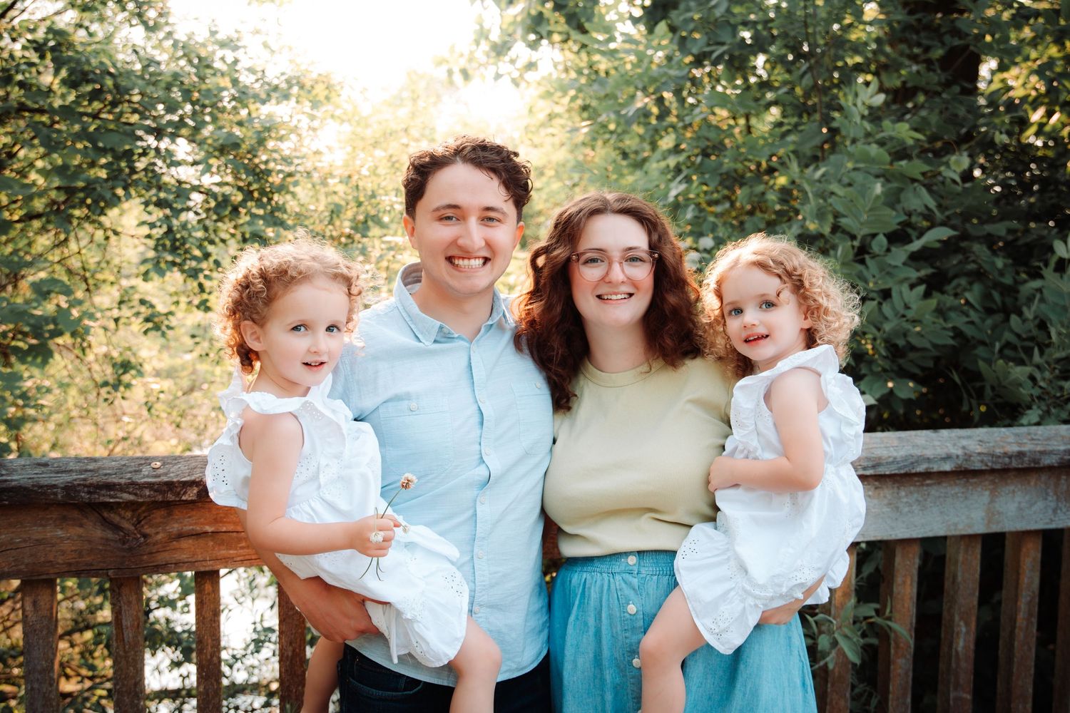 Family of four poses together on wooden deck with lush green foliage in background.