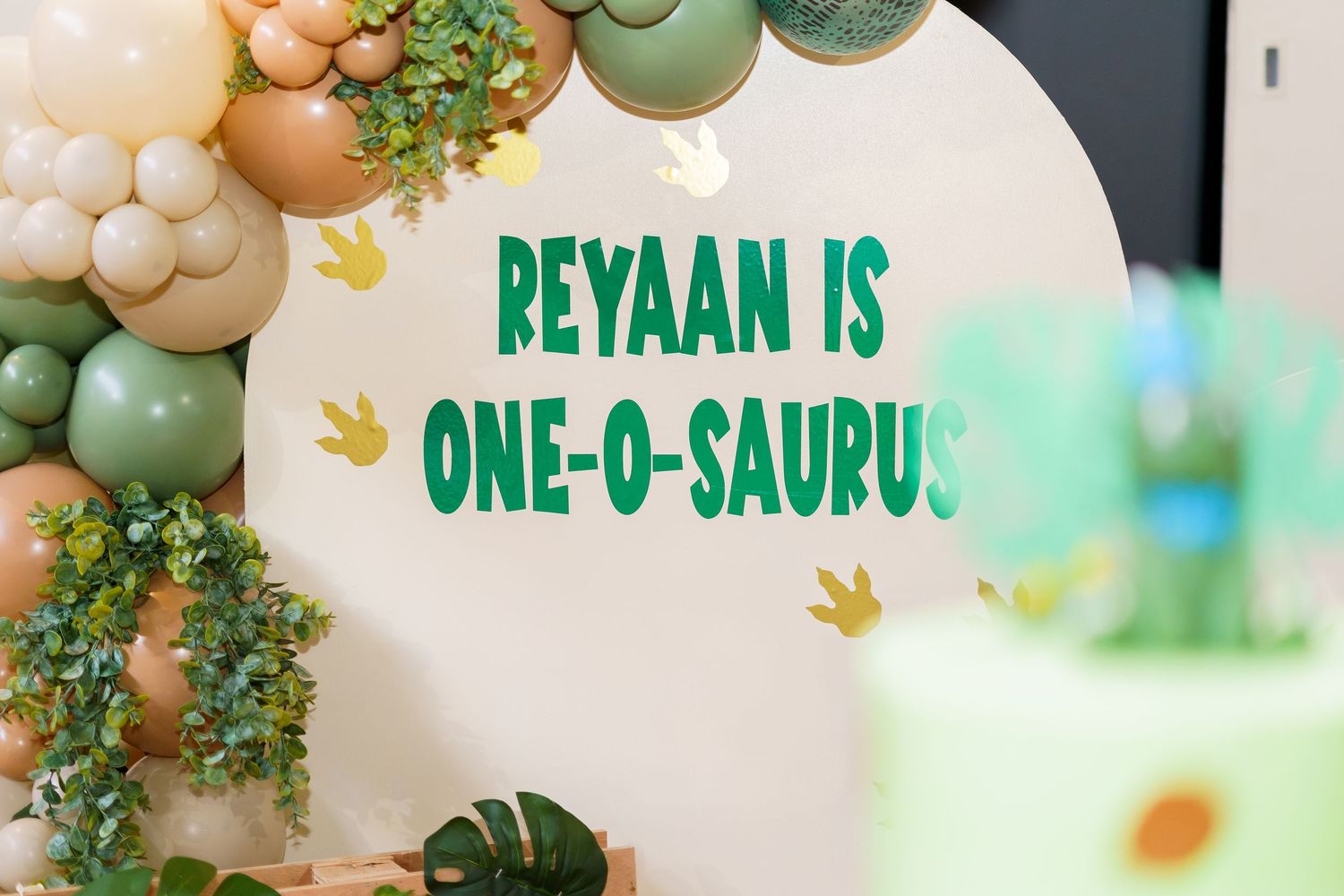 Birthday party decorations with green text reading 'REYAAN IS ONE-O-SAURUS' on white background with balloon garland.