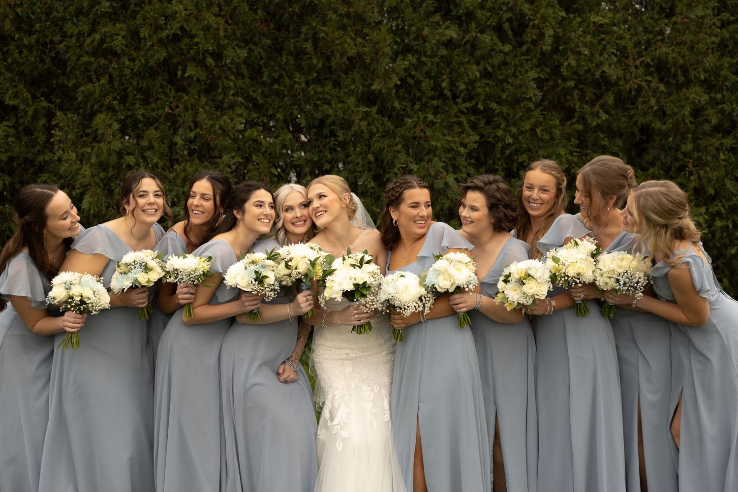 Wedding party wearing light gray dresses hold white bouquets while sharing a joyful moment together at outdoor venue.