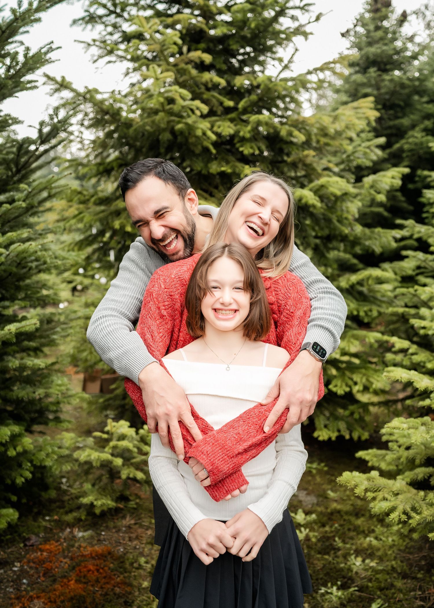 A happy family shares a warm embrace among evergreen trees on a bright day.